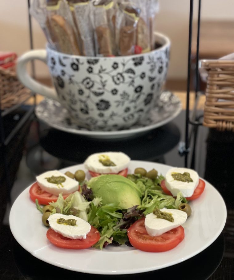 A caprese-style plate of mozzarella, tomato, avocado and capers with a floral cappuccino cup behind
